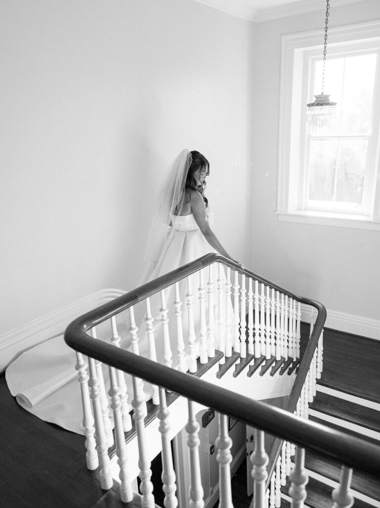 Black and white aerial view looking down the grand staircase at McAlister-Leftwich Greensboro NC with Grace from behind in a cathedral veil and ballgown train, hand resting on the wood railing — Raleigh wedding photographer