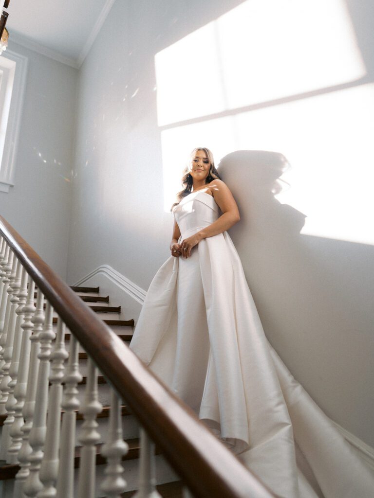Dramatic low-angle bridal portrait of Grace on the grand staircase at McAlister-Leftwich Greensboro NC, sunlight streaming through the window with lens flares, strapless ballgown train cascading down the wood steps — Raleigh wedding photographer