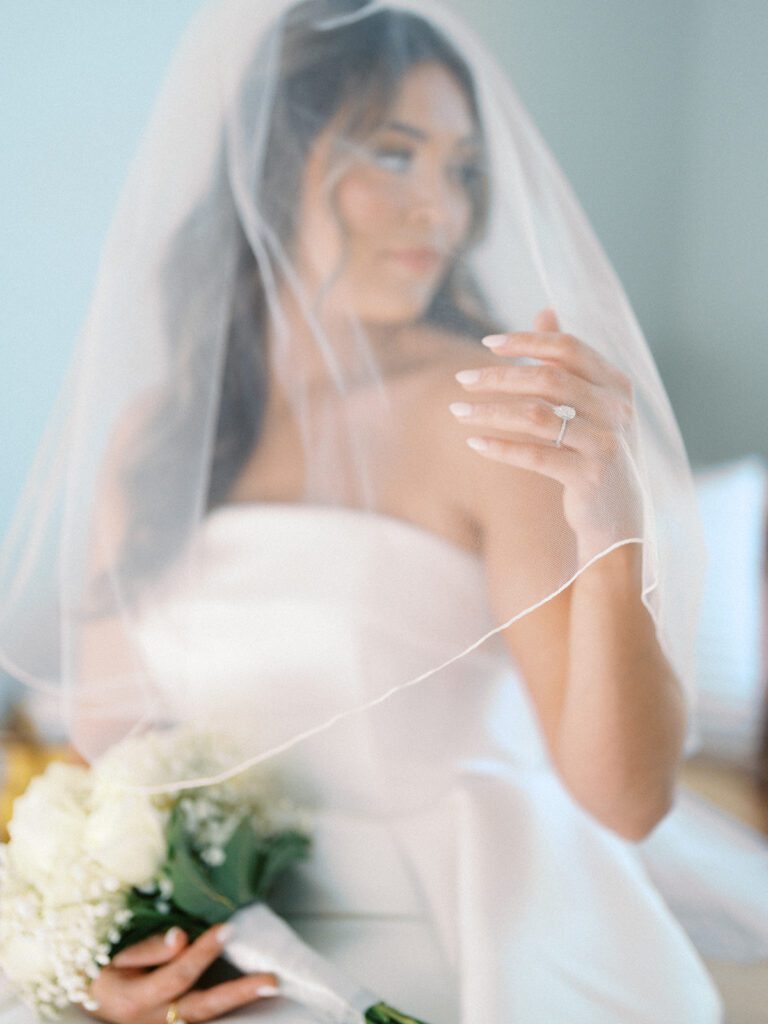 Romantic soft-focus bridal portrait shot through the cathedral veil at McAlister-Leftwich Greensboro NC, Grace's hand with her diamond engagement ring resting on her chest and white rose bouquet in the foreground — Raleigh wedding photographer