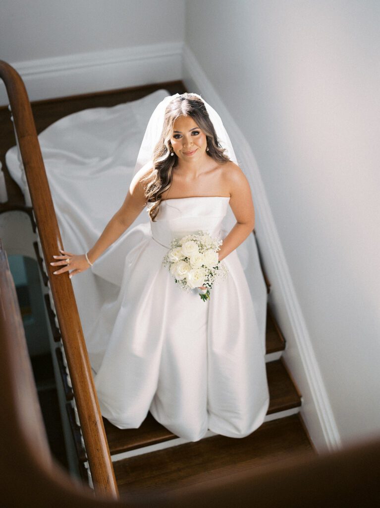 Grace smiling up at the camera from the grand staircase at McAlister-Leftwich in Greensboro NC, wearing a cathedral veil and holding a white rose and baby's breath bouquet, strapless ballgown train on the stairs — Raleigh wedding photographer