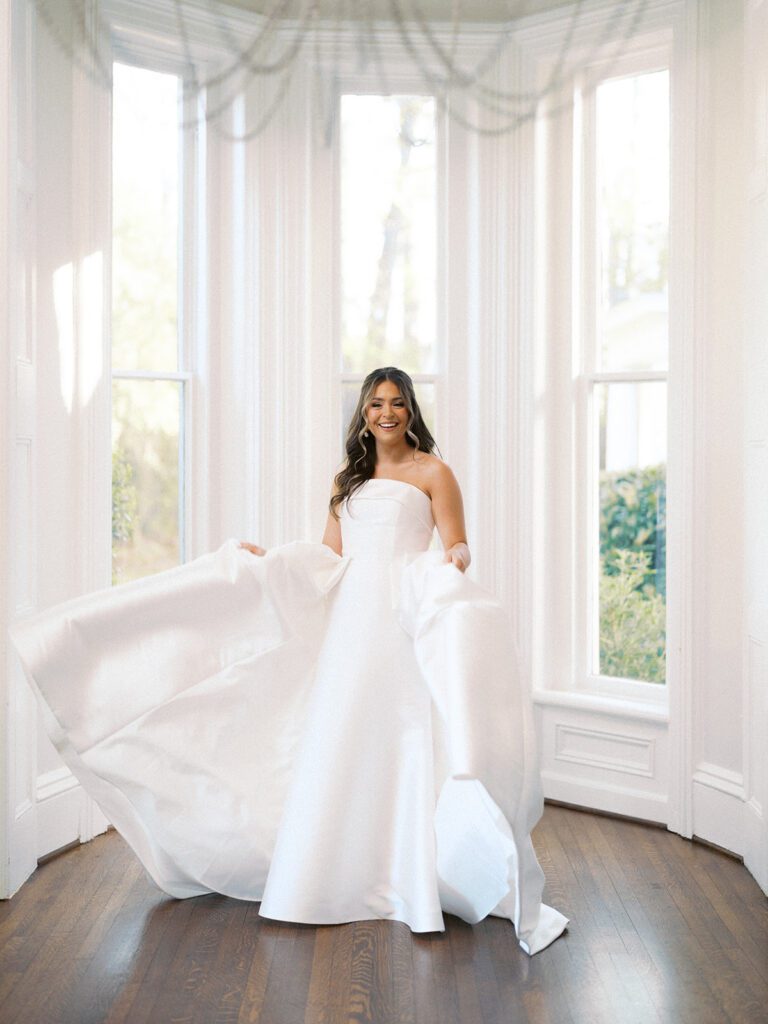 Grace joyfully twirling in her strapless white ballgown in the floor-to-ceiling bay window sunroom at McAlister-Leftwich Greensboro NC, skirt flying out as she laughs — Raleigh wedding photographer