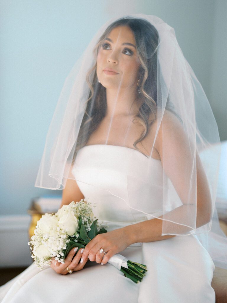 Bride seated with a cathedral veil draped over her face, gazing upward while holding a white rose bouquet with her diamond engagement ring visible in the blue room at McAlister-Leftwich Greensboro NC — Raleigh wedding photographer