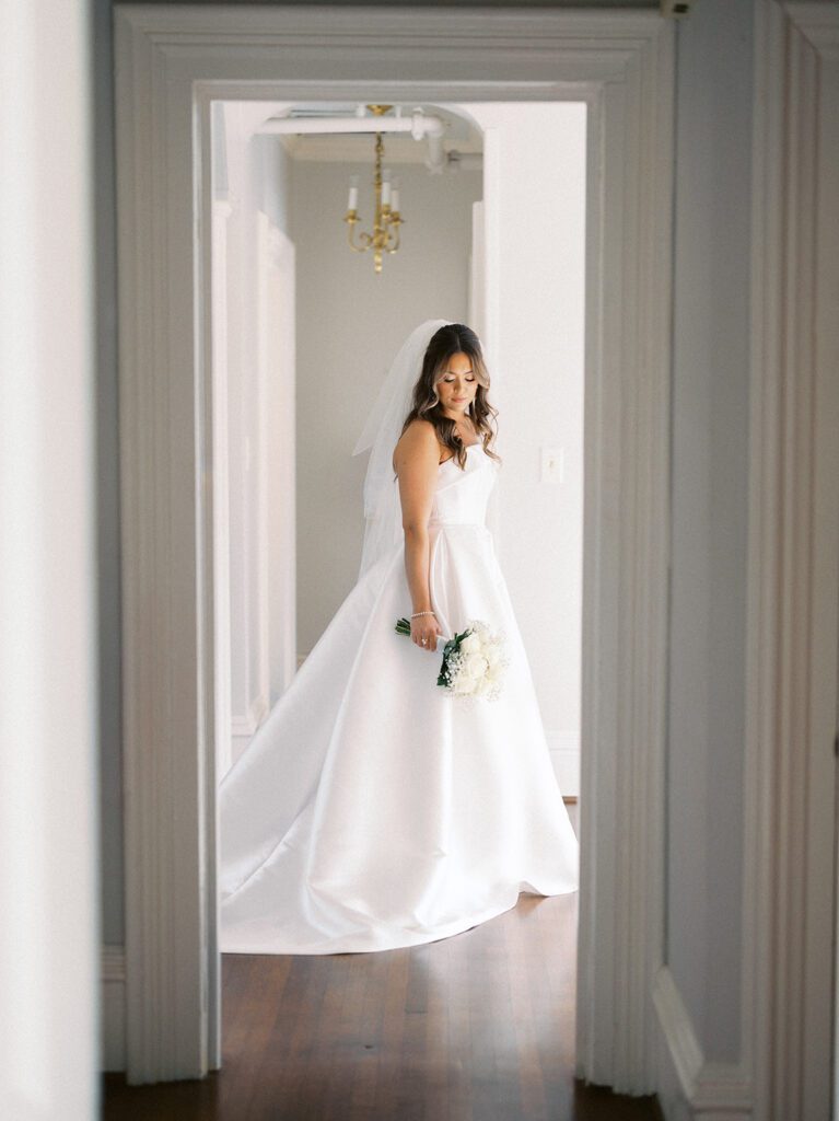 Full-length bridal portrait of Grace in a cathedral veil and strapless ballgown beautifully framed by a white doorway arch at McAlister-Leftwich Greensboro NC, looking down at her white rose bouquet — Raleigh wedding photographer