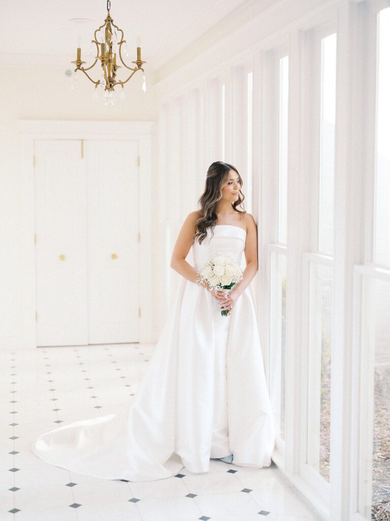 Full-length bridal portrait of Grace in a strapless white ballgown with cathedral train holding a white rose and baby's breath bouquet while gazing out the window in the chandelier hallway at McAlister-Leftwich Greensboro NC — Raleigh wedding photographer